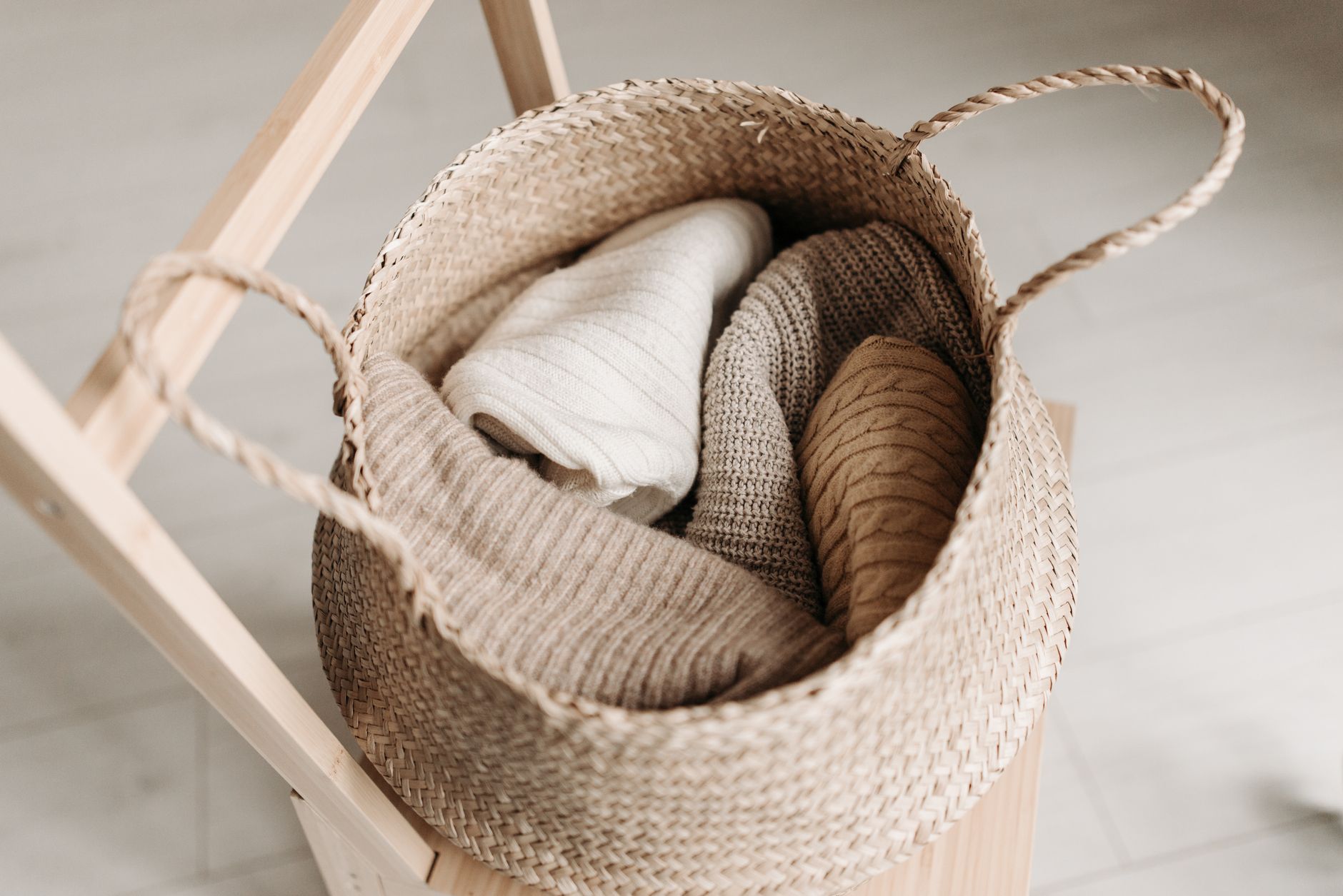 Folded knitted garments in a woven basket on a wooden chair.